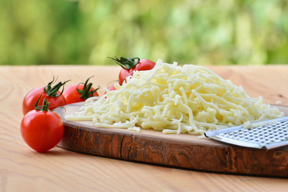 Grated pizza cheese on a cutting board and some tomatoes on the side